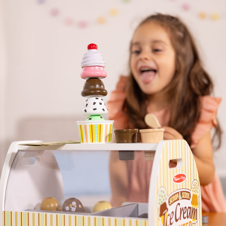 A kid playing with The Melissa & Doug Wooden Scoop and Serve Ice Cream Counter With Play Food and Accessories (28 Pcs)