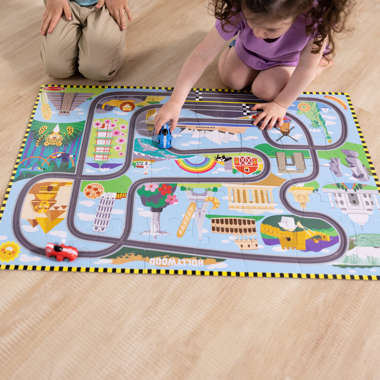 A kid playing with The Melissa & Doug Race Around the World Tracks Cardboard Jigsaw Floor Puzzle and Wind-Up Vehicles – 48 Pieces, for Boys and Girls 3+