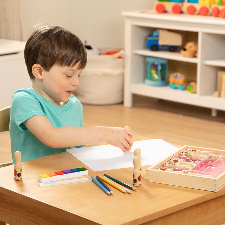 A kid playing with The Melissa & Doug Deluxe Happy Handle Stamp Set With 10 Stamps, 5 Colored Pencils, and 6-Color Washable Ink Pad