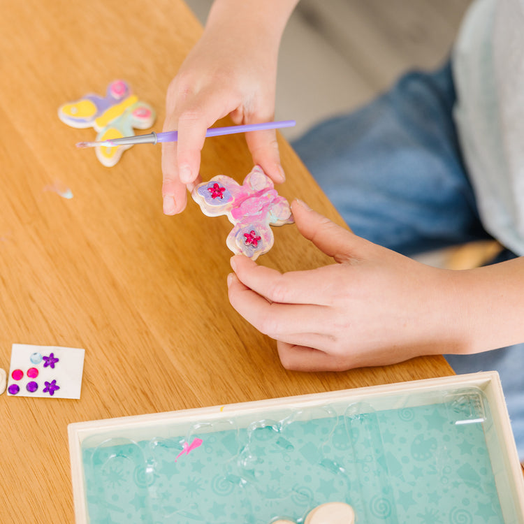 A kid playing with The Melissa & Doug Created by Me! Wooden Butterfly Magnets Craft Kit (4 Designs, 4 Paints, Stickers, Glitter Glue)