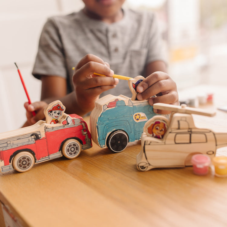 A kid playing with The Melissa & Doug PAW Patrol Wooden Vehicles Craft Kit - 3 Decorate Your Own Vehicles, 3 Play Figures