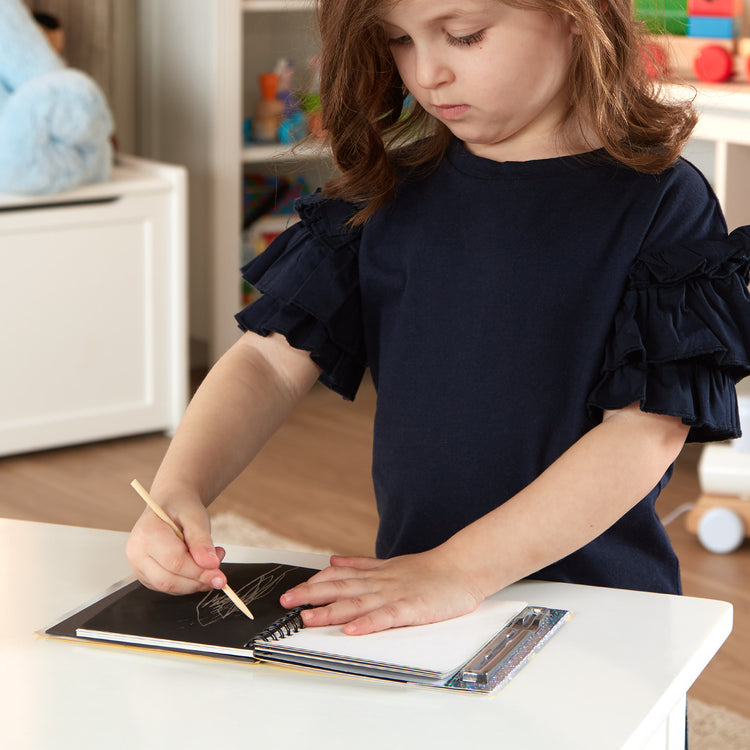 A kid playing with The Melissa & Doug Scratch Art Doodle Pad With 16 Scratch-Art Boards and Wooden Stylus