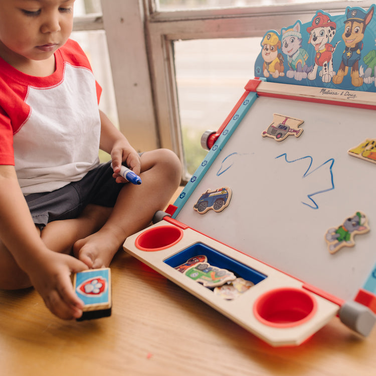 A kid playing with The Melissa & Doug PAW Patrol Wooden Double-Sided Tabletop Art Center Easel (33 Pieces)