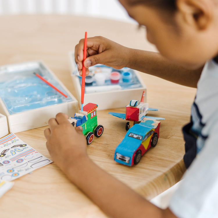 A kid playing with The Melissa & Doug Decorate-Your-Own Wooden Craft Kits 3-Pack - Plane, Train, and Race Car