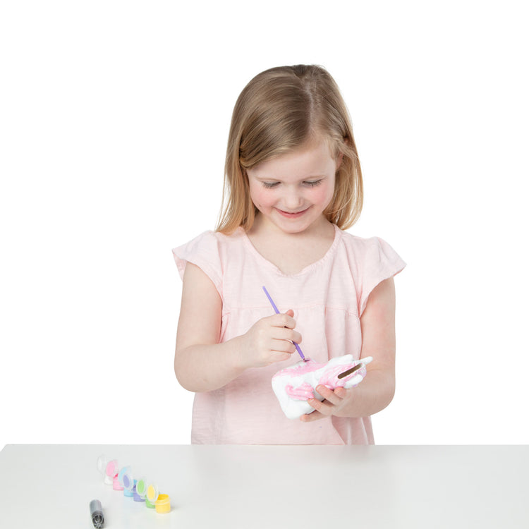 A child on white background with The Melissa & Doug Created by Me! Decorate-Your-Own Unicorn Bank Craft Kit With 6 Pots of Paint, Glitter Glue, Paintbrush