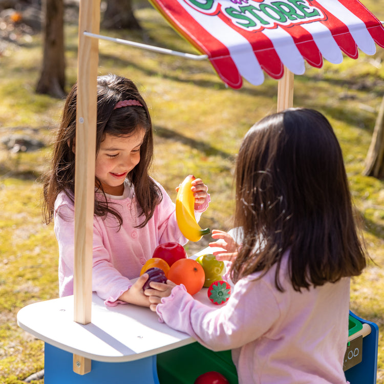 A kid playing with The Melissa & Doug Wooden Grocery Store and Lemonade Stand Activity Center - Reversible Awning, 9 Bins, 9 Chalkboards