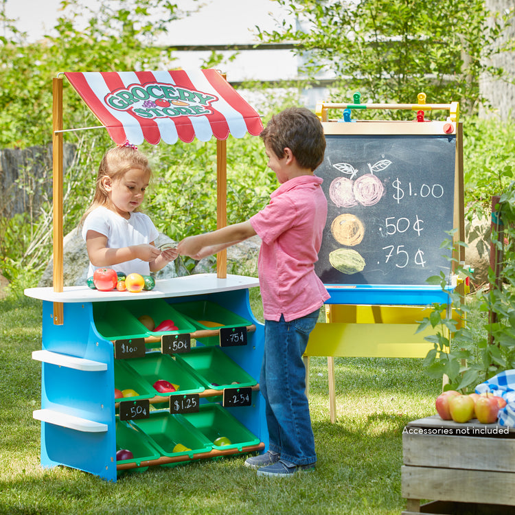 A kid playing with The Melissa & Doug Wooden Grocery Store and Lemonade Stand Activity Center - Reversible Awning, 9 Bins, 9 Chalkboards