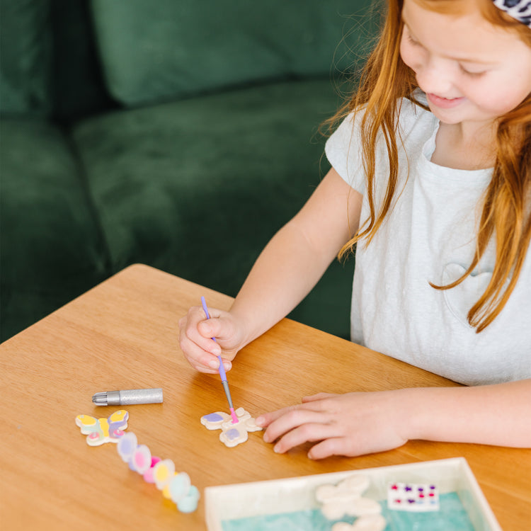 A kid playing with The Melissa & Doug Created by Me! Wooden Butterfly Magnets Craft Kit (4 Designs, 4 Paints, Stickers, Glitter Glue)