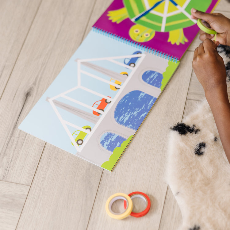 A kid playing with The Melissa & Doug Tape Activity Book: 4 Rolls of Easy-Tear Tape and 20 Reusable Scenes