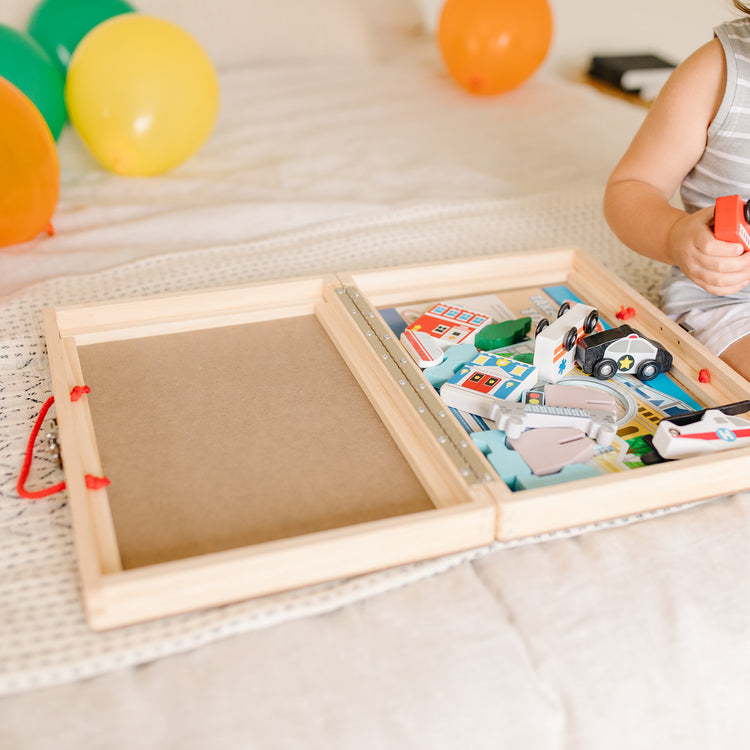 A kid playing with The Melissa & Doug 18-Piece Wooden Take-Along Tabletop Town, 4 Rescue Vehicles, Play Pieces, Bridge