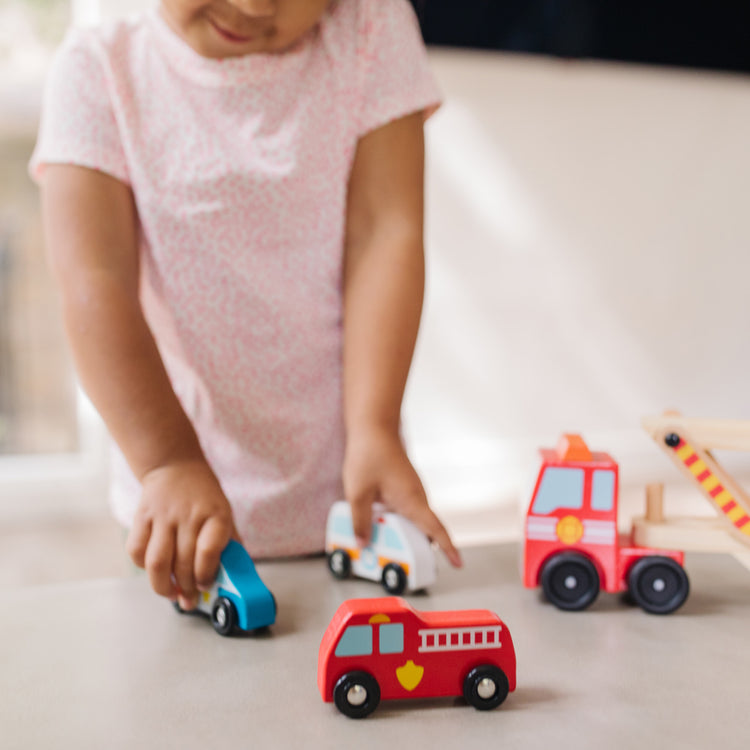 A kid playing with The Melissa & Doug Wooden Emergency Vehicle Carrier Truck With 1 Truck and 4 Rescue Vehicles