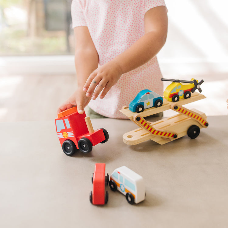 A kid playing with The Melissa & Doug Wooden Emergency Vehicle Carrier Truck With 1 Truck and 4 Rescue Vehicles