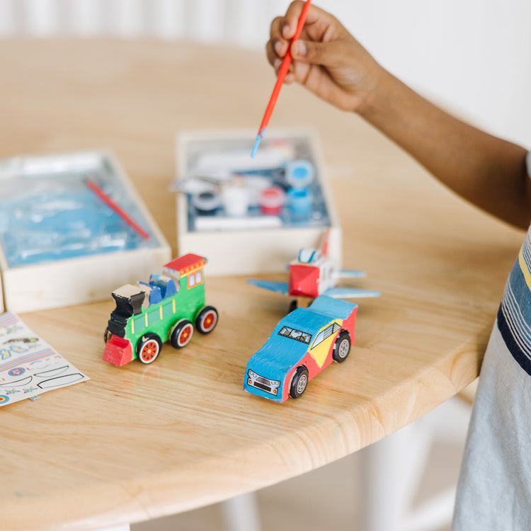 A kid playing with The Melissa & Doug Decorate-Your-Own Wooden Craft Kits 3-Pack - Plane, Train, and Race Car