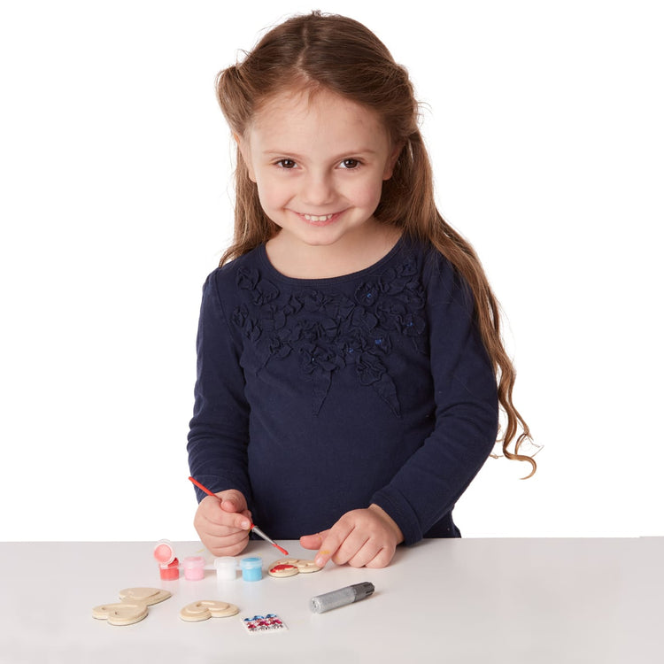 A child on white background with the Melissa & Doug Created by Me! Wooden Heart Magnets Craft Kit (4 Designs, 4 Paints, Stickers, Glitter Glue)