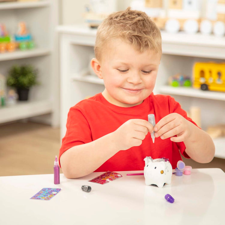 A kid playing with the Melissa & Doug Created by Me! Piggy Bank Craft Kit with 4 Pots of Paint, Brush, Glitter, Stickers