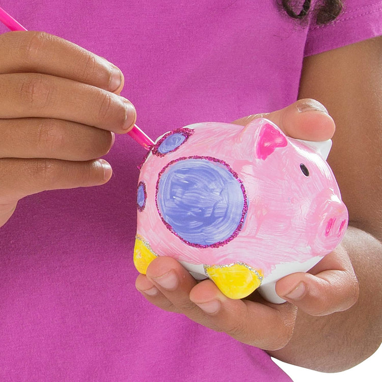 A child on white background with the Melissa & Doug Created by Me! Piggy Bank Craft Kit with 4 Pots of Paint, Brush, Glitter, Stickers